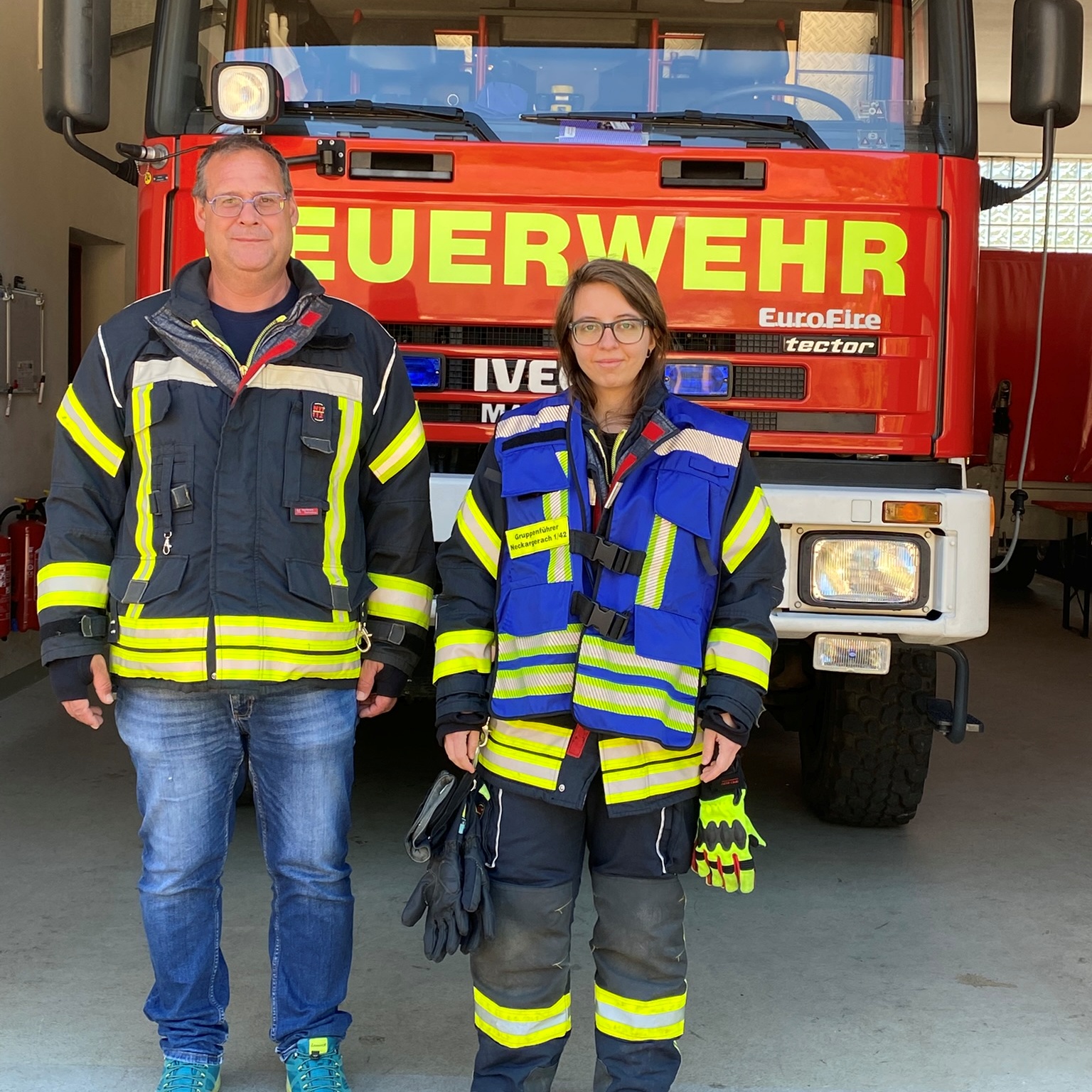 Ein Mann und eine Frau stehen mit Feuerwehrkleidung vor einem Feuerwehrauto. Die Frau trägt eine blaue Weste.