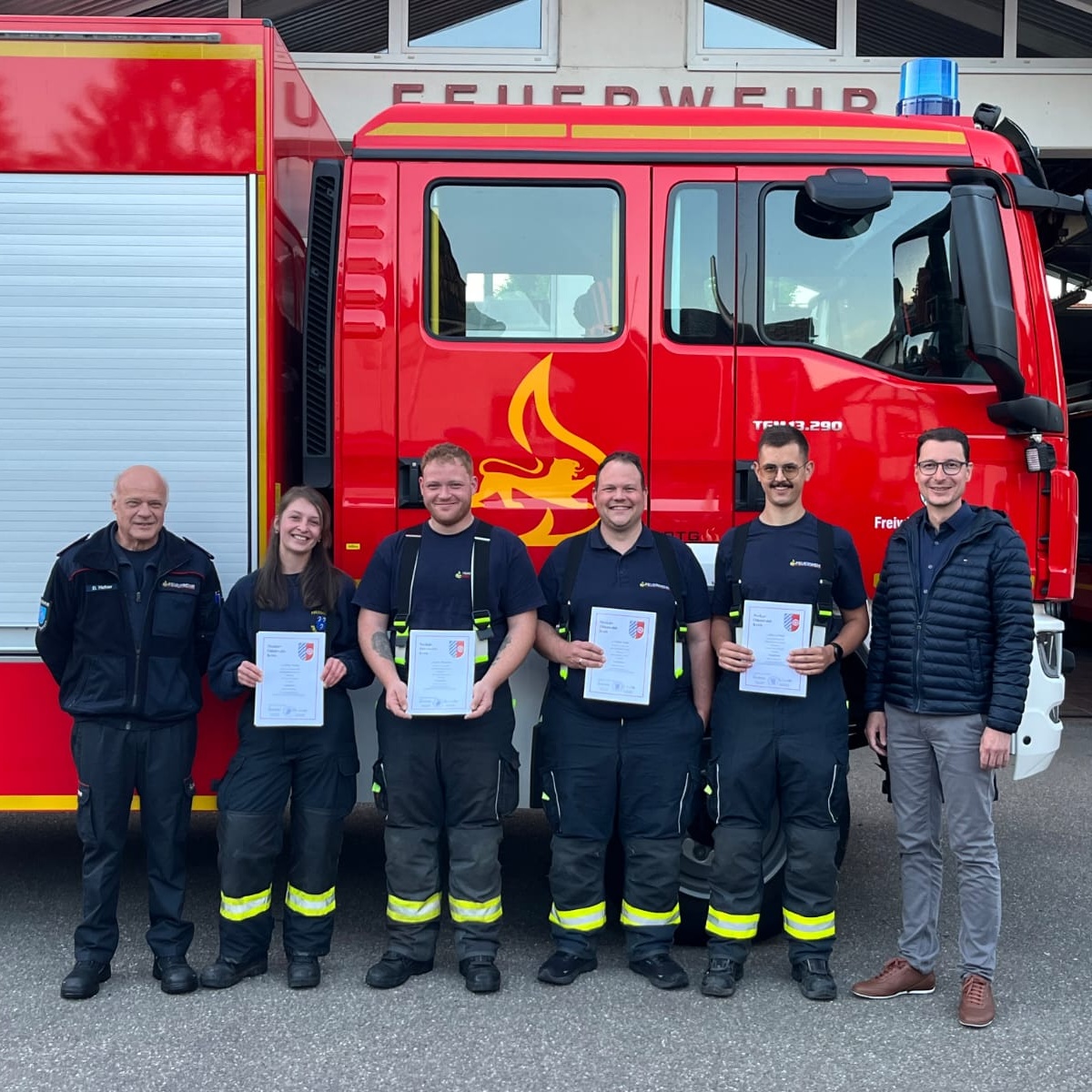 Sechs Personen stehen vor einem Feuerwehrauto. Die vier in der Mitte tragen Feuerwehr-Schutzhosen und halten Urkunden in der Hand.