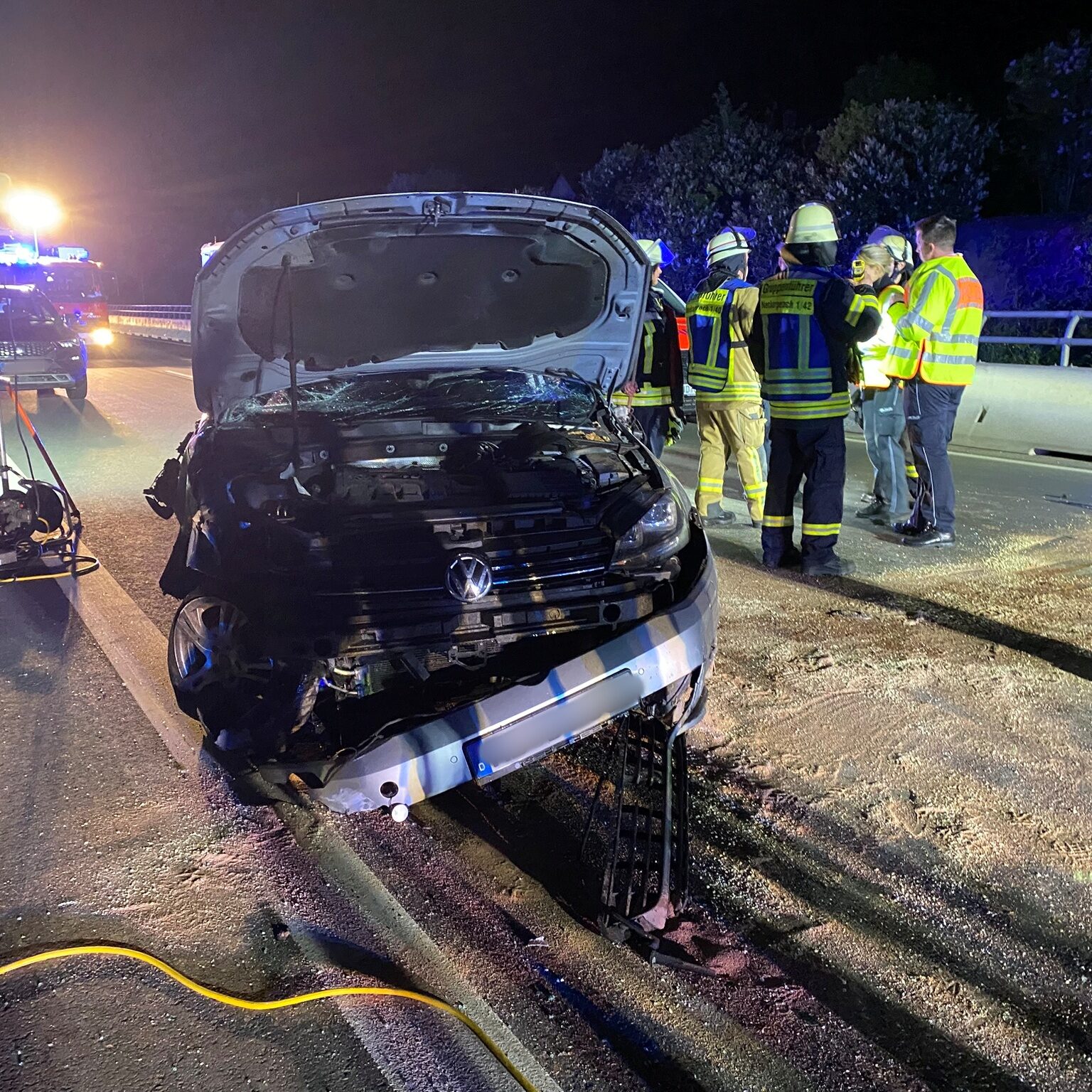 Ein Auto, die Motorhaube ist hochgehoben und die Hälfte des Autos ist kaputt. Im Hintergrund mehrere Feuerwehrleute und Polizisten.
