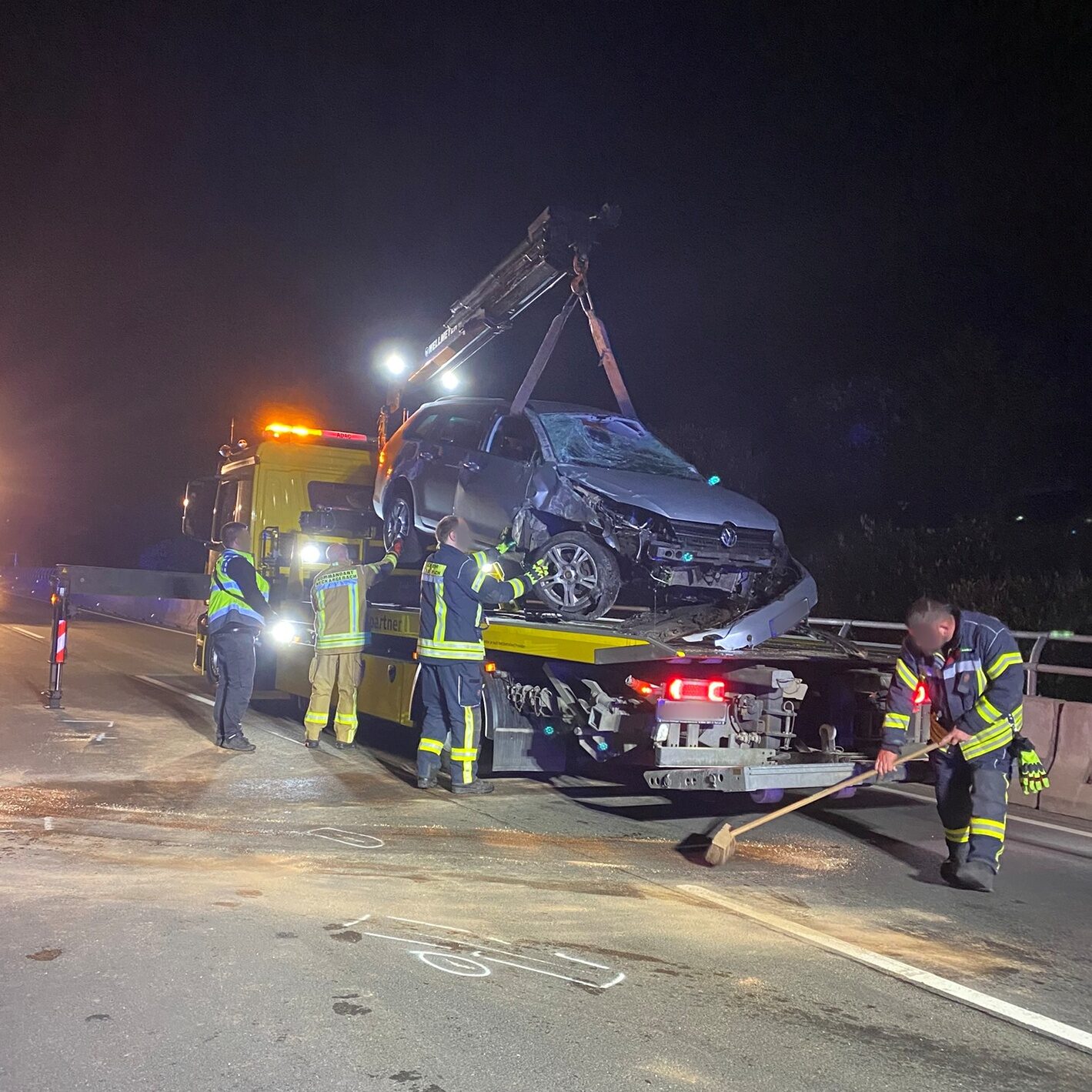 Ein kaputtes Auto wird von einem Kran auf einen Abschlepper gehoben. Drei Feuerwehrleute stehen dabei.