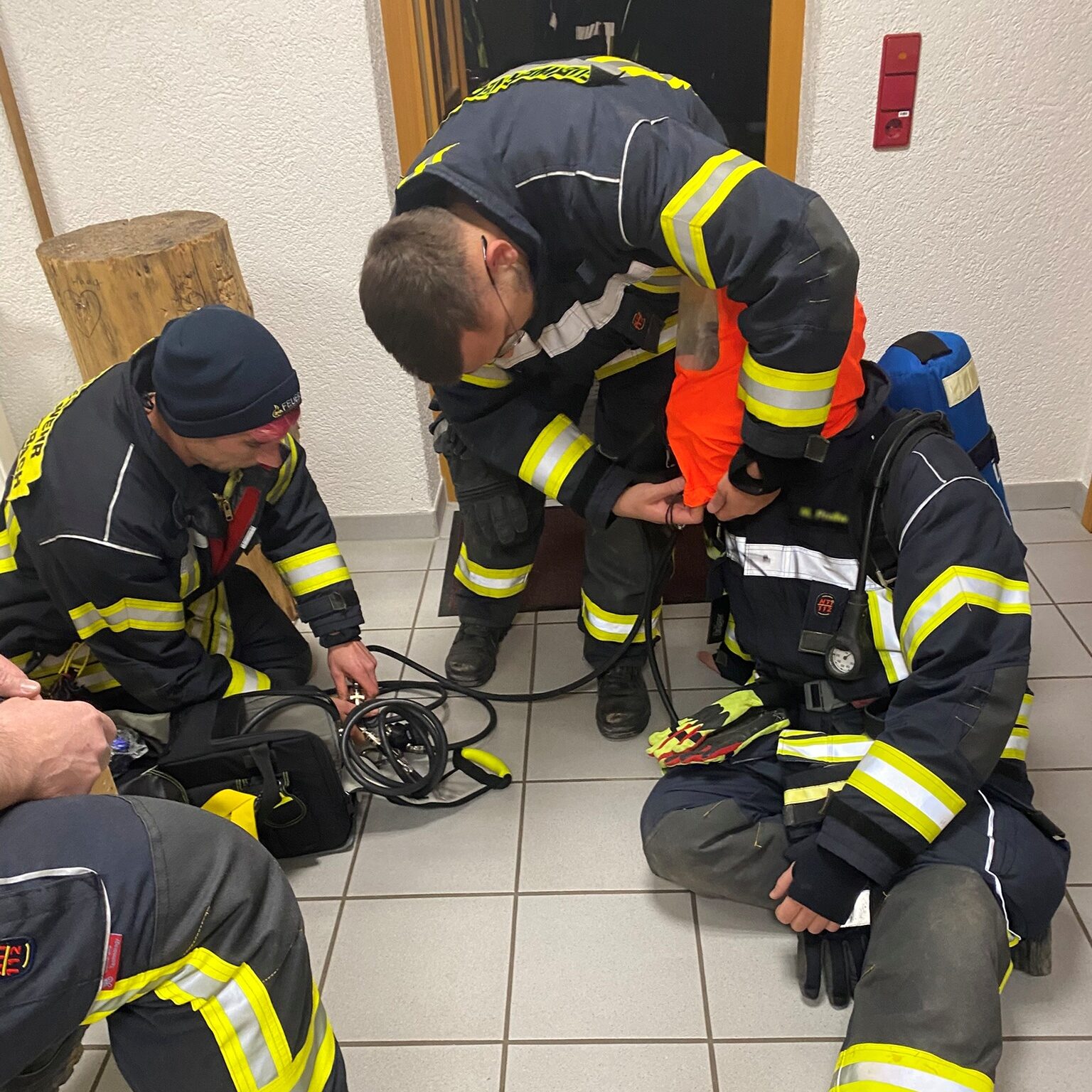 Ein Feuerwehrmann sitzt auf dem Boden und bekommt von einem anderen Feuerwehrmann eine rote Maske übergezogen.