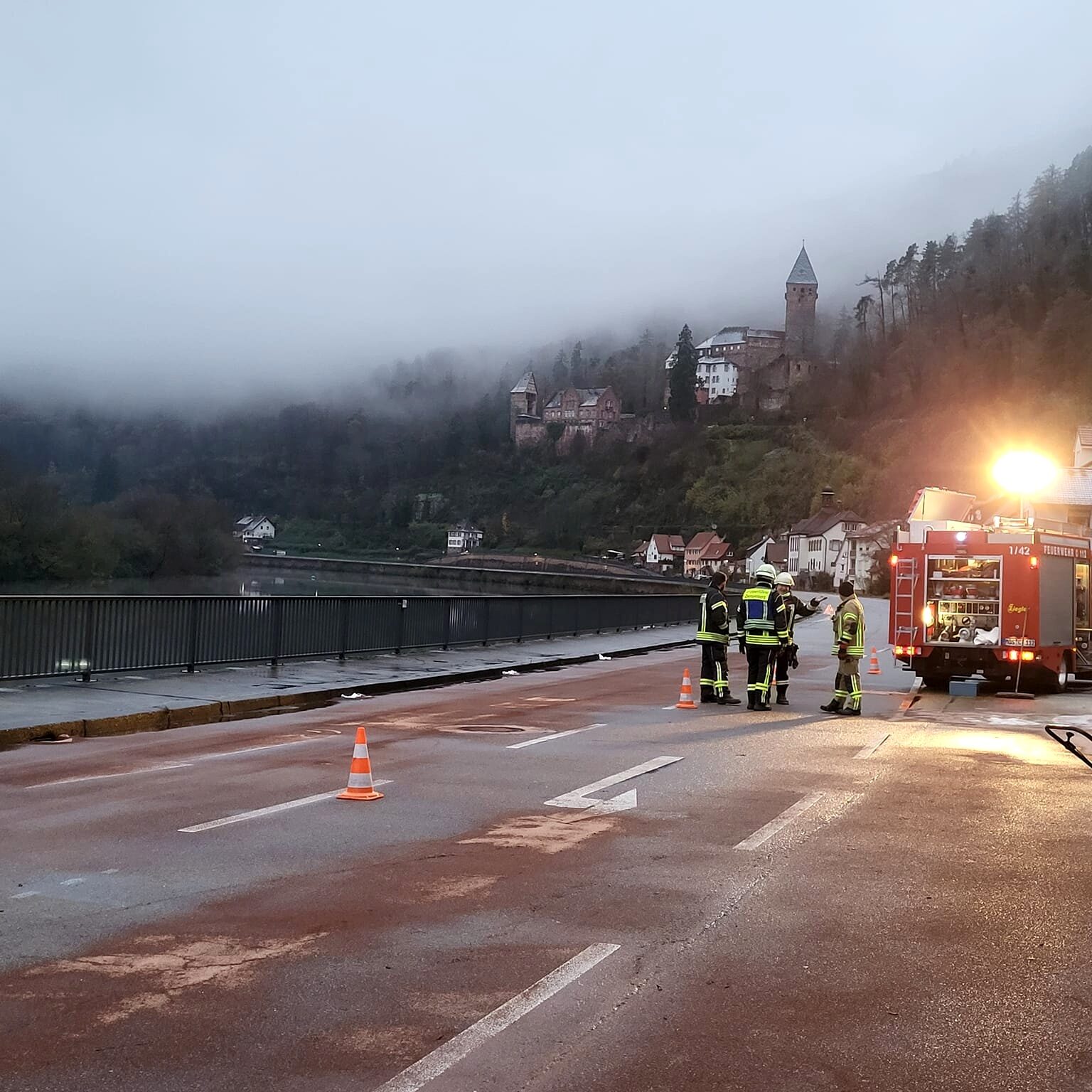 Eine gesperrte Straße, auf der braunes Pulver liegt. Feuerwehrleute stehen bei einem Feuerwehrauto. Im Hintergrund sieht man ein Schloss im Nebel.