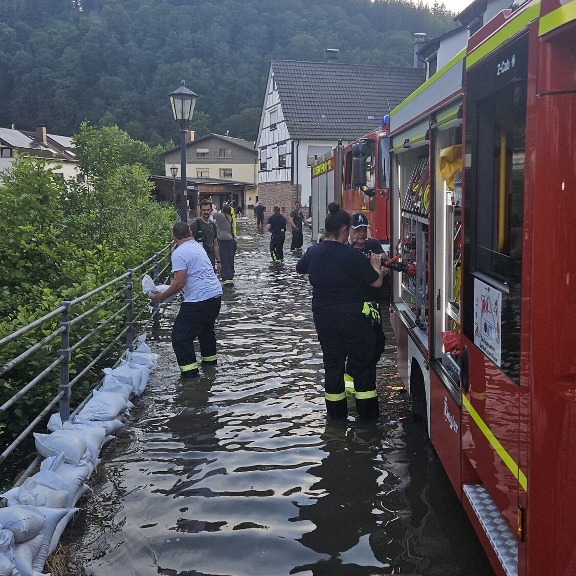 Eine Straße ist mit Wasser überschwemmt. Rote Feuerwehrautos stehen im Wasser. Menschen legen Sandsäcke an einen Zaun.