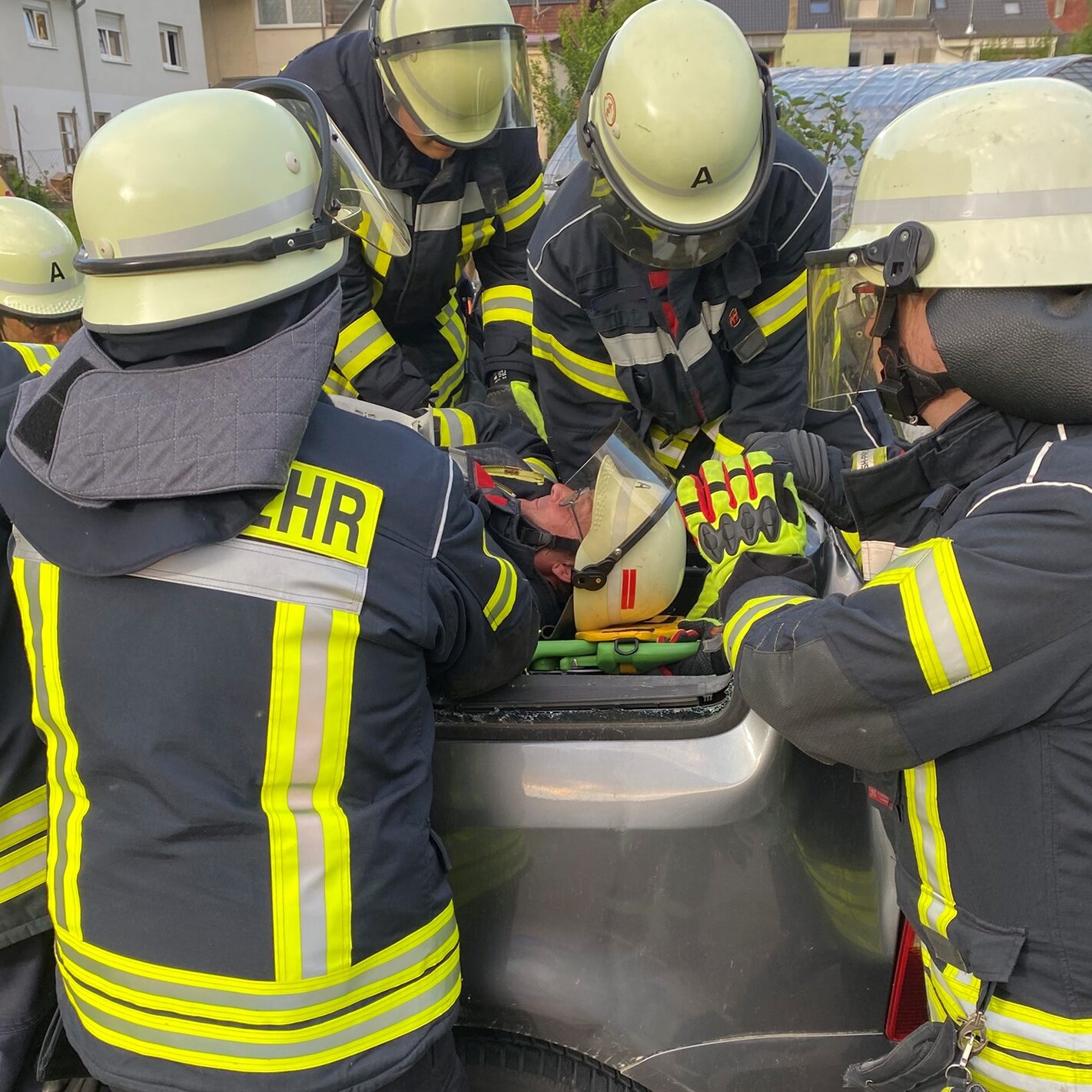Fünf Männer in Feuerwehrkleidung stehen an einem Auto, in der mitte liegt ein Mann auf einer Trage mit Feuerwehrkleidung.