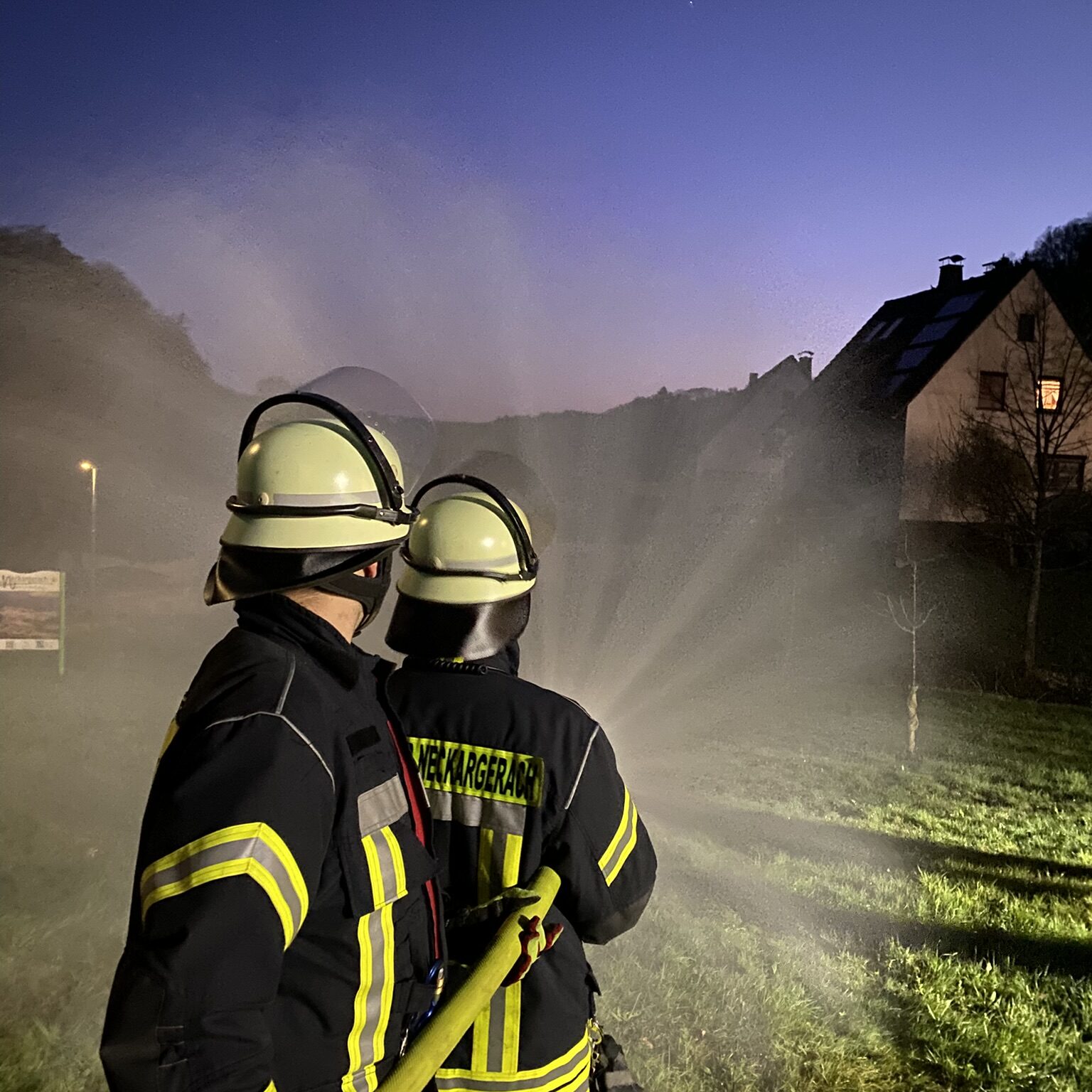 Zwei Feuerwehrmänner halten einen Schlauch, aus dem Wasser in einem weiten Strahl herauskommt auf eine Wiese.