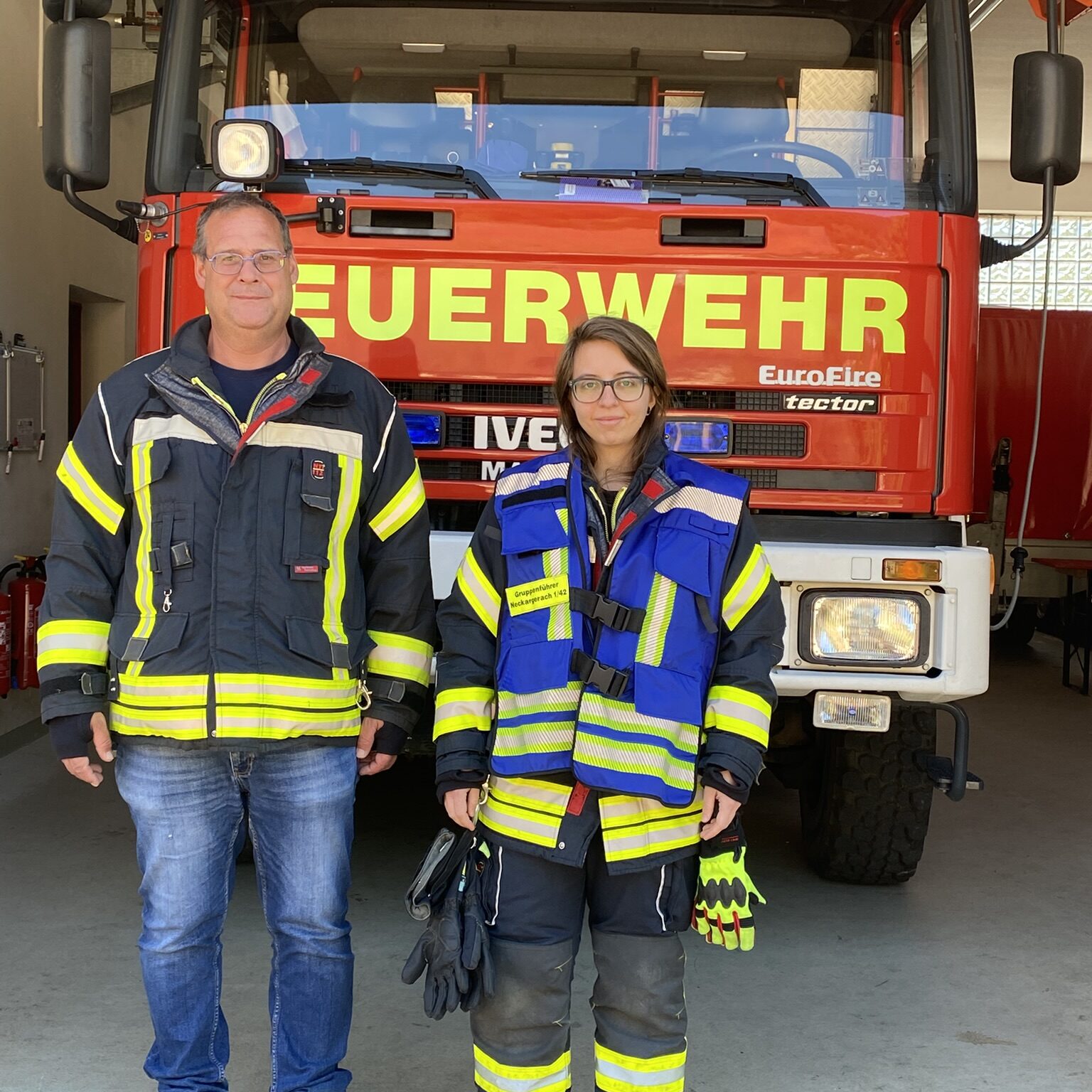 Ein Mann und eine Frau stehen mit Feuerwehrkleidung vor einem Feuerwehrauto. Die Frau trägt eine blaue Weste.