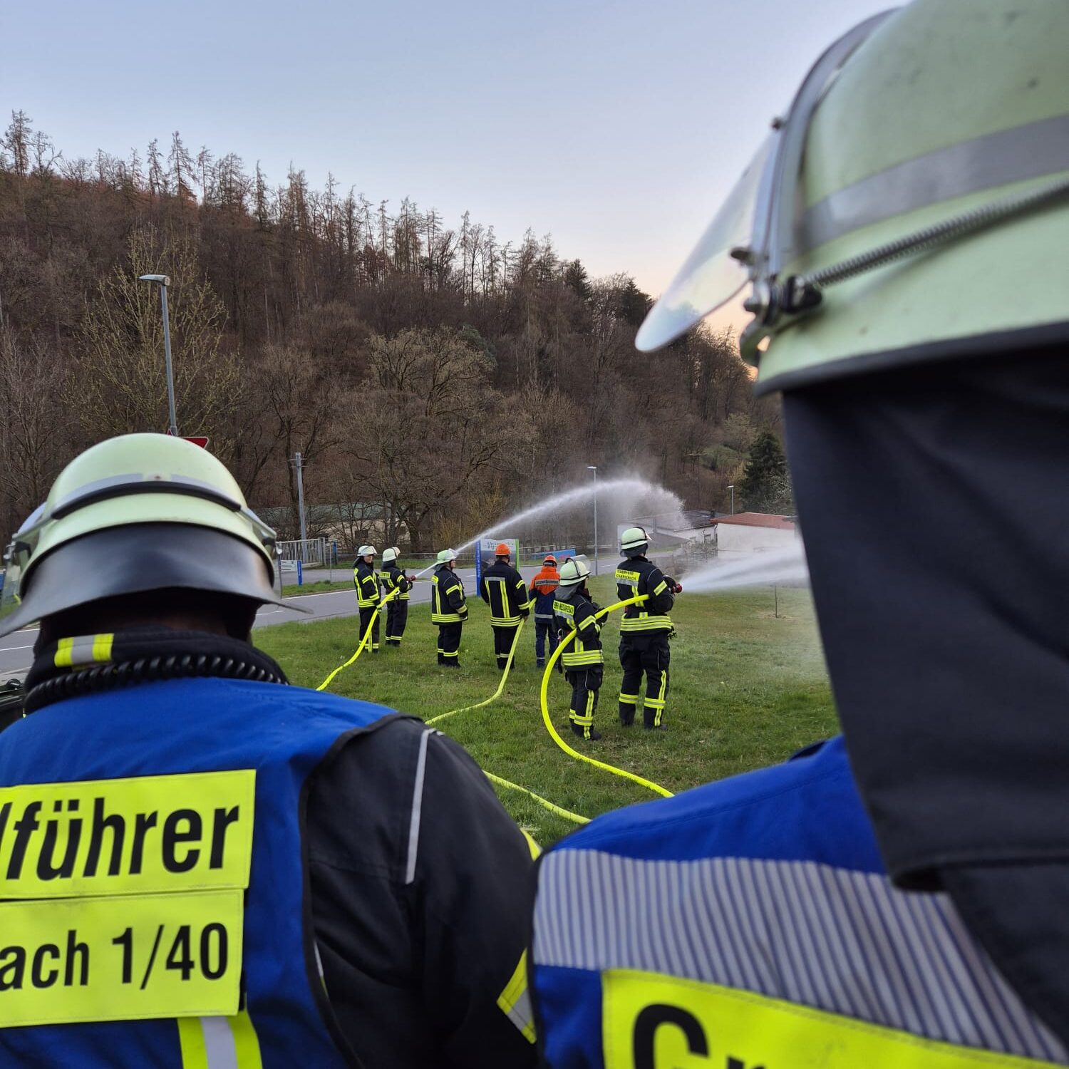 Blick über die Schulte von zwei Feuerwehrleuten mit blauer Weste auf andere Feuerwehrleute, die Wasser auf eine Wiese spritzen.