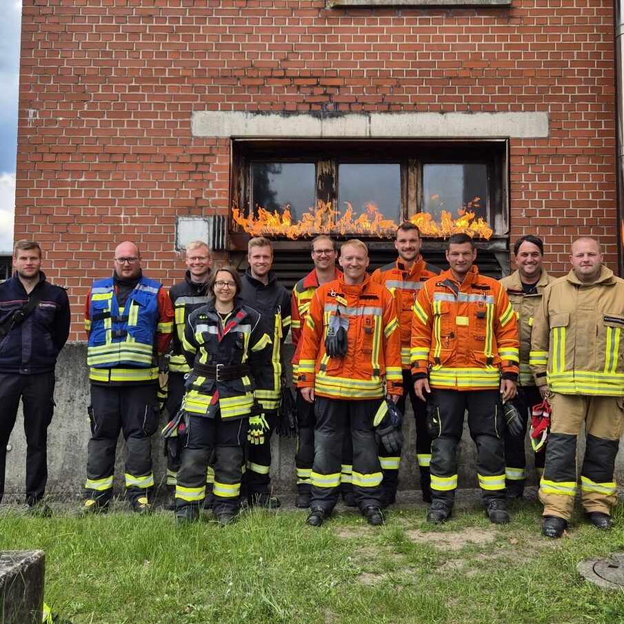 Eine Gruppe von Feuerwehrleuten in verschiedenfarbiger Einsatzkleidung steht vor einem Backsteinhaus, am Fenster brennt ein kleines Feuer.