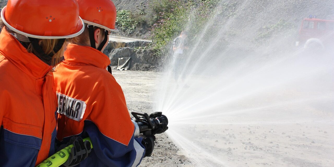 youth-fire-brigade-4665039_1280 Zwei Kinder mit orangenem Helm und orange und blauer Jacke halten einen Schlauch, aus dem Wasser spritzt.
